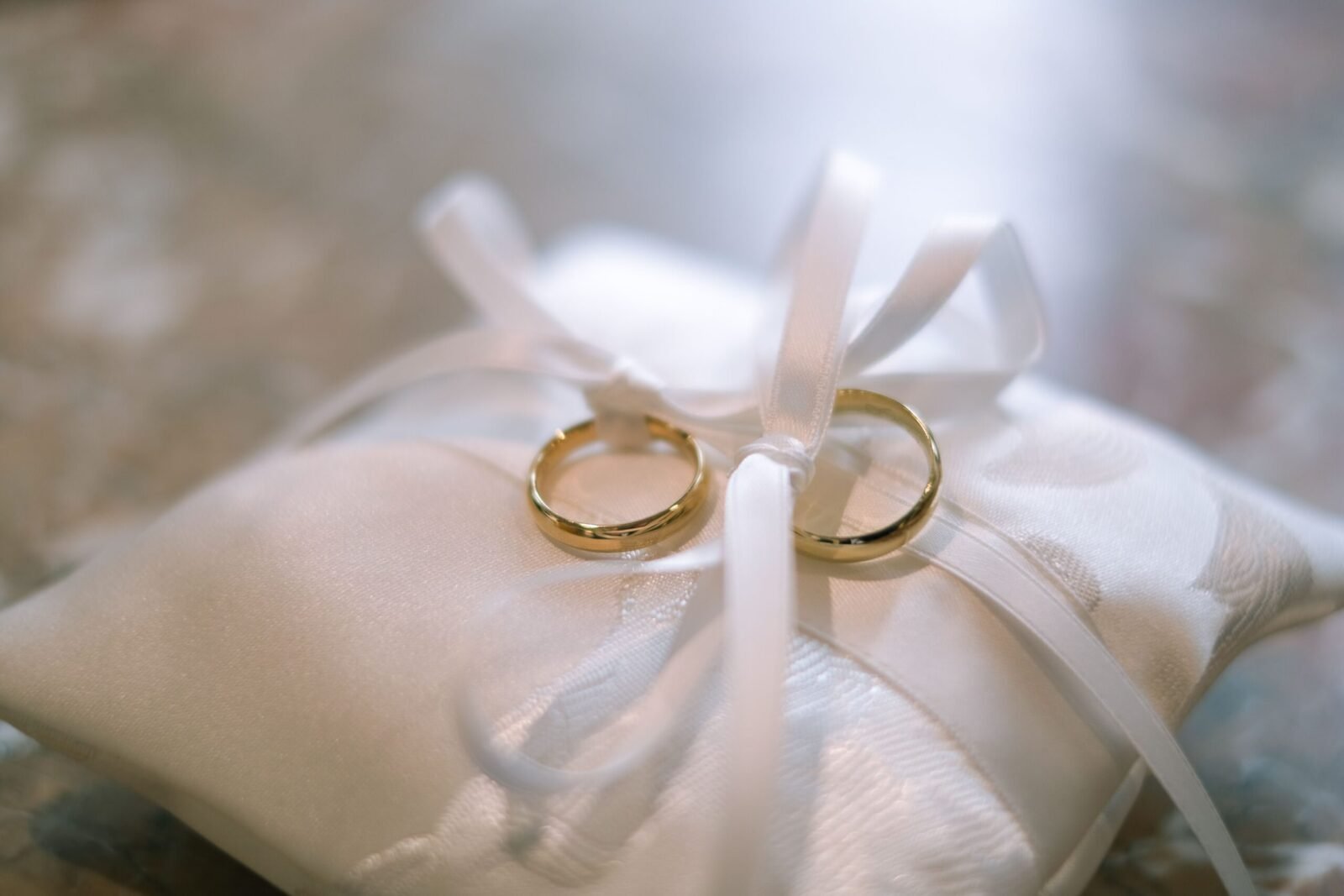 Close-up of two gold wedding rings on a satin pillow tied with ribbons, symbolizing marriage.